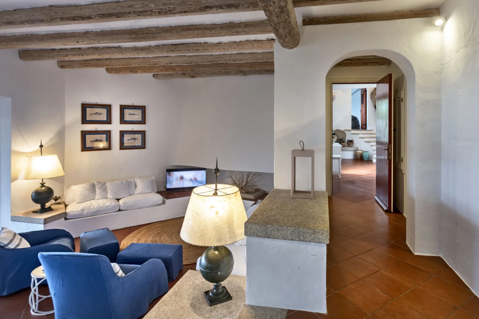 Cozy living room with blue chairs, a white sofa, and rustic wooden beams, featuring a lamp and a television, leading to an arched doorway into another room. | Only on SopranoVillas
