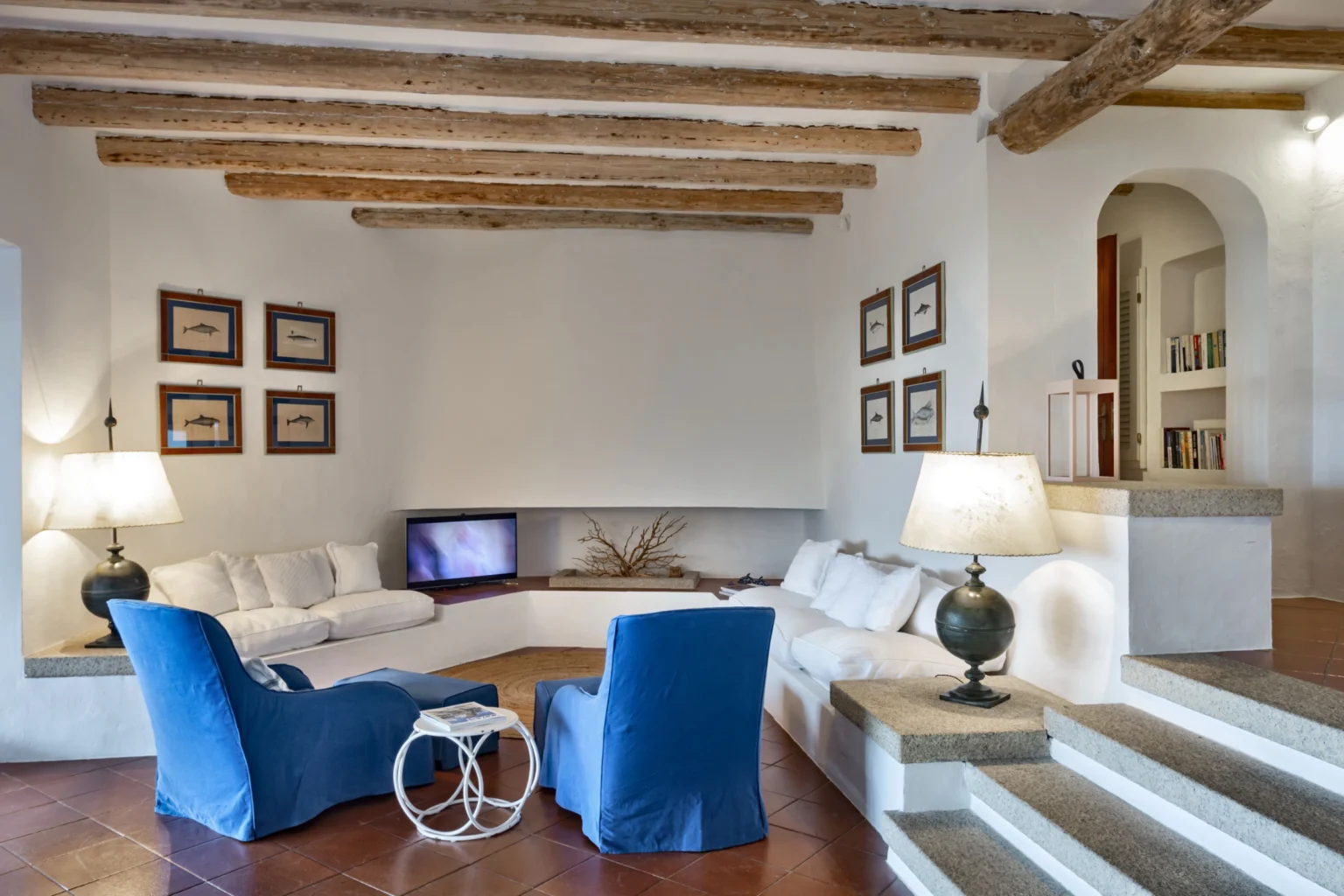 Modern living room with exposed wooden beams, featuring a cozy white sectional sofa, two blue chairs, a small round coffee table, and wall art. The space is well-lit with lamps and has a warm, inviting atmosphere. | Only on SopranoVillas