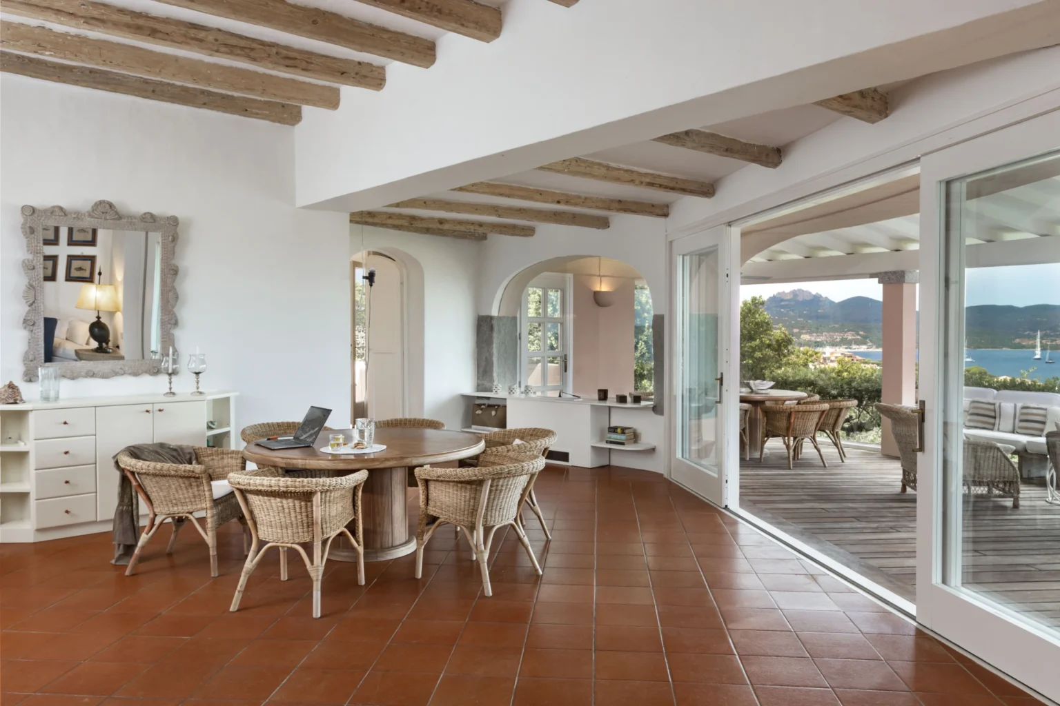 Bright and airy dining area featuring a round wooden table surrounded by wicker chairs, with large windows offering views of the outdoor patio and scenic landscape. The room showcases a combination of white walls and rustic wooden beams, enhancing the modern coastal aesthetic. | Only on SopranoVillas