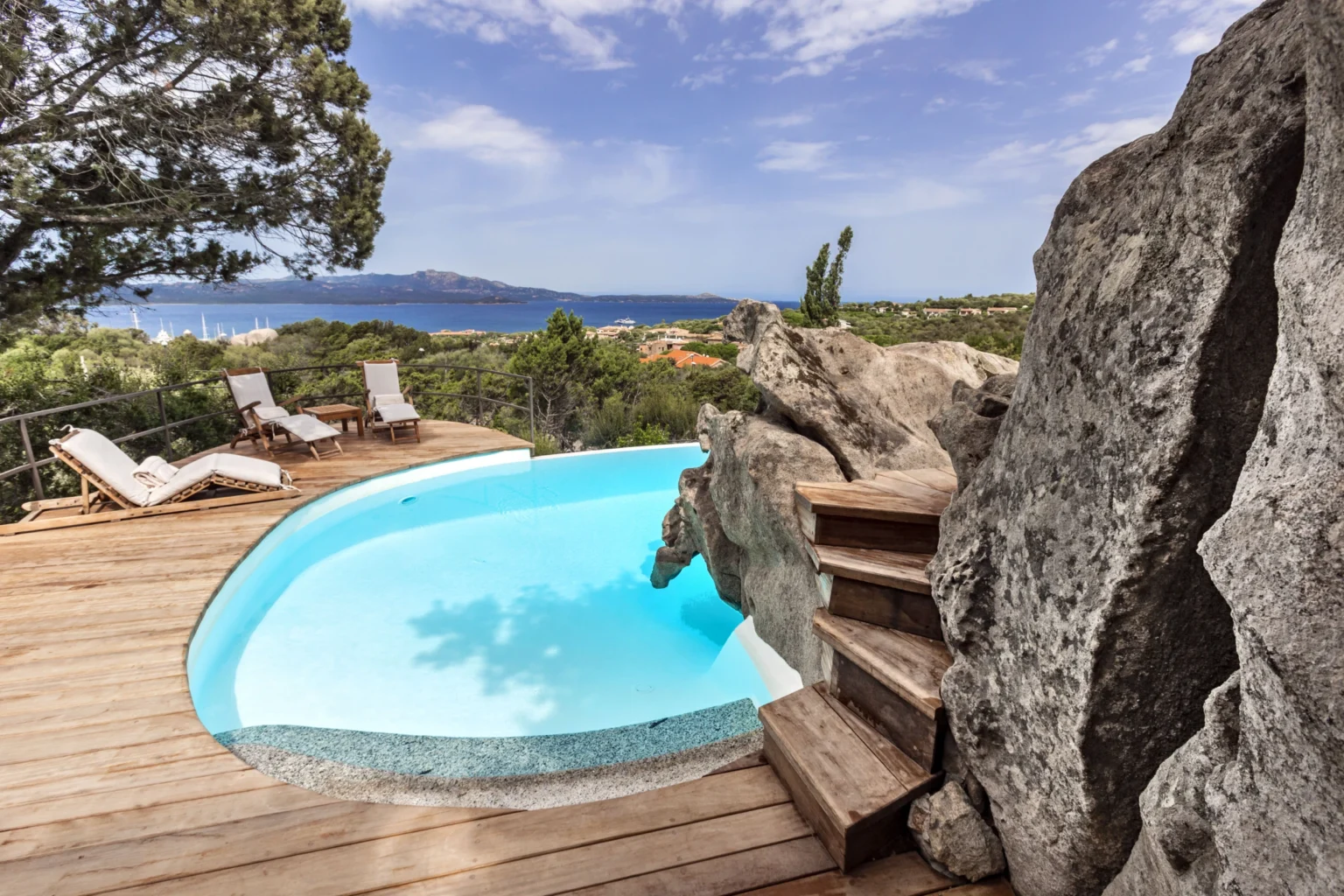 Scenic outdoor pool with wooden deck and natural rock formation, overlooking a coastal landscape and blue sky. Perfect for relaxation and luxury retreats. | Only on SopranoVillas