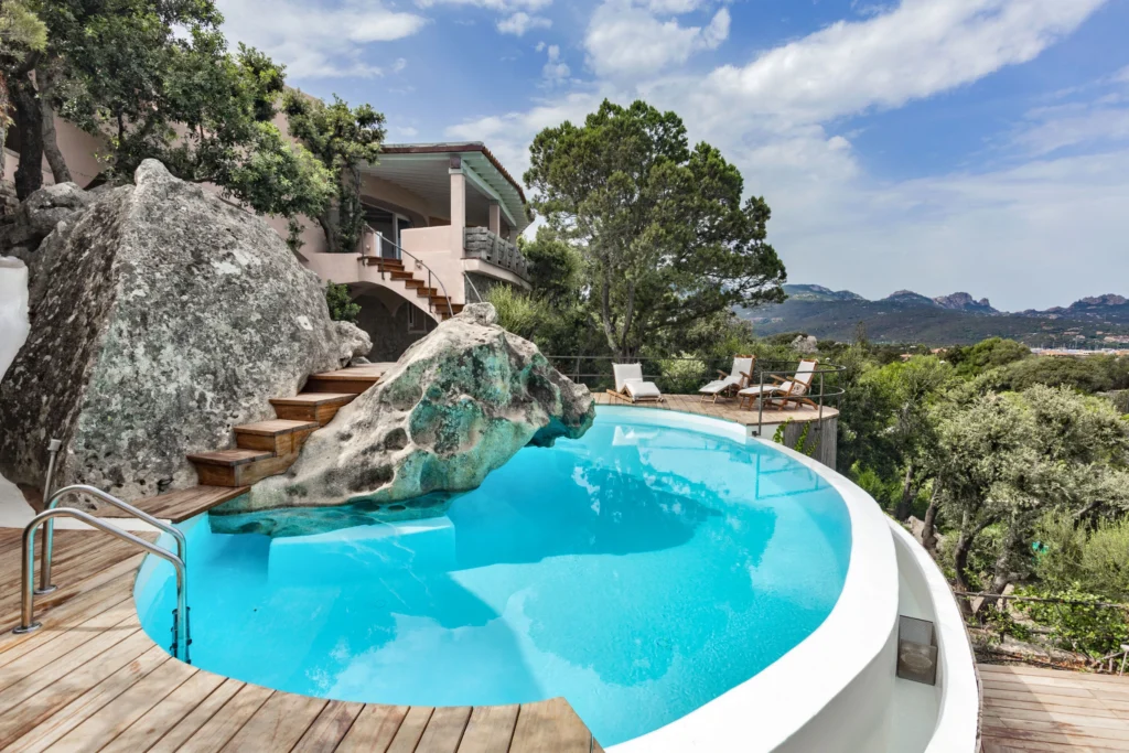 Luxury outdoor pool with natural rock feature, surrounded by lush greenery and modern architecture, under a clear blue sky. Ideal for relaxation and scenic views. | Only on SopranoVillas