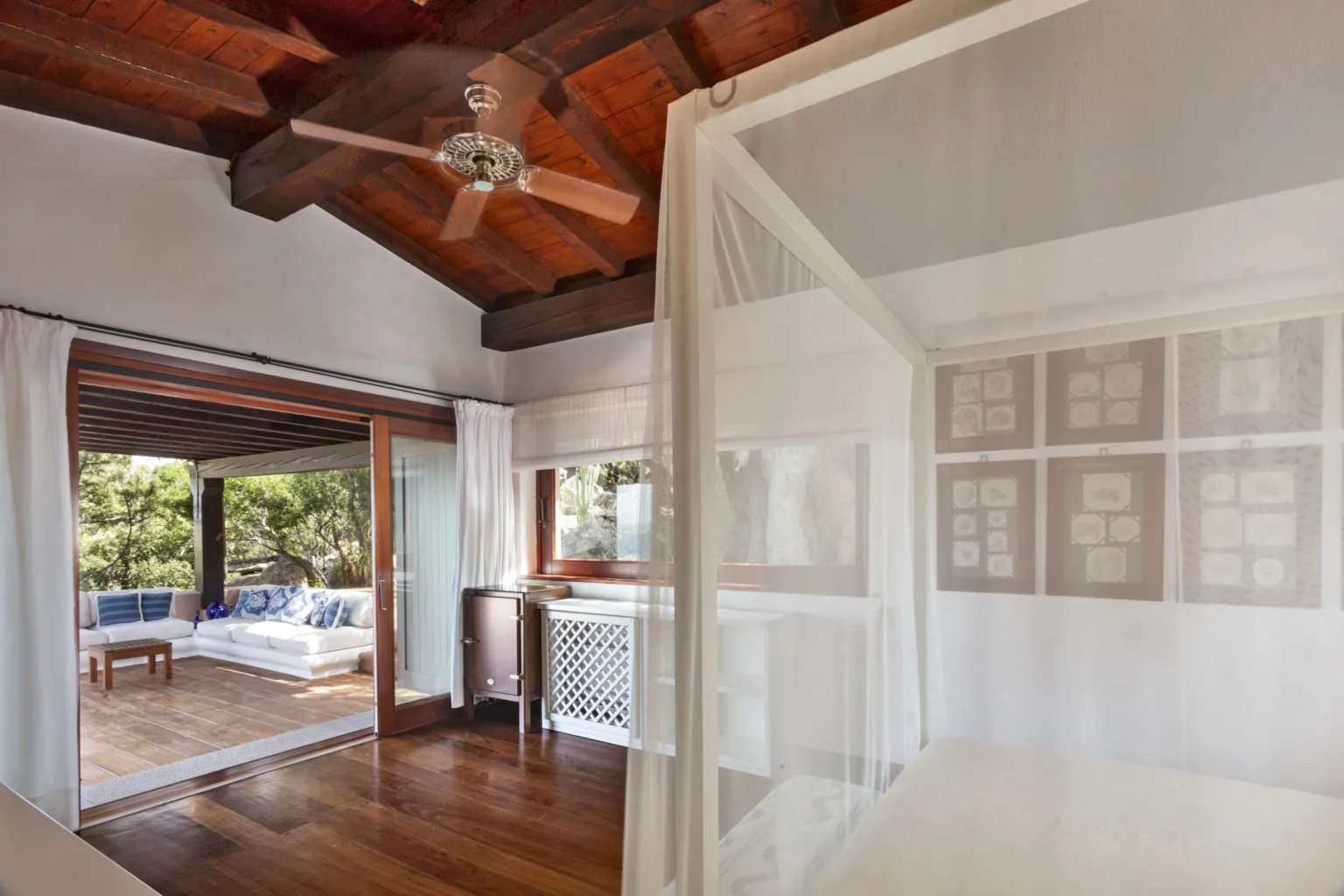 Bright and airy bedroom with wooden ceiling beams, featuring a cozy bed draped with sheer curtains, and a view of a sunny outdoor lounge area through large glass doors. | Only on SopranoVillas