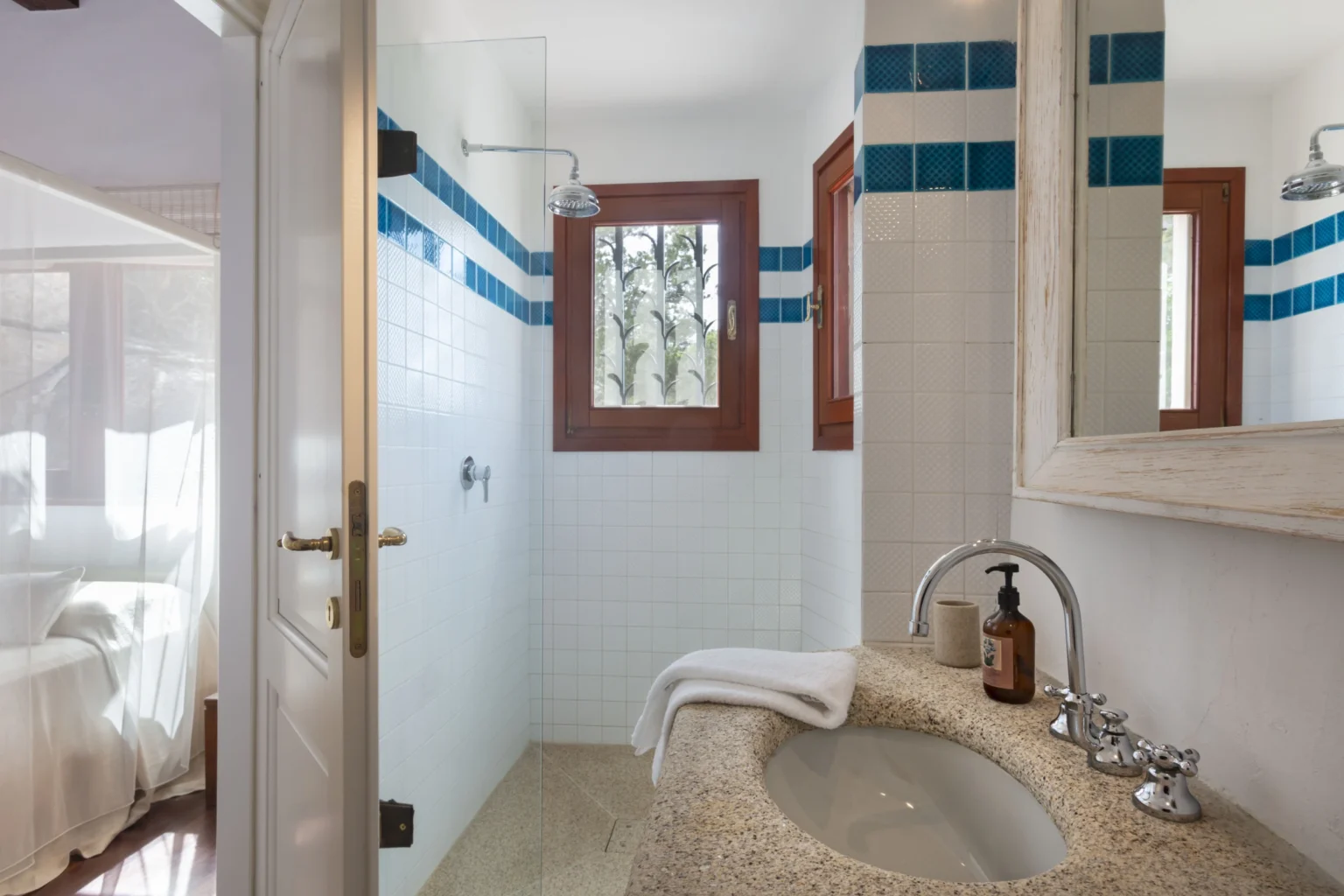 Modern bathroom with a stylish sink, natural stone countertop, and blue accent tiles. Features a large mirror, shower area, and wooden window frames, creating a bright and inviting space. | Only on SopranoVillas