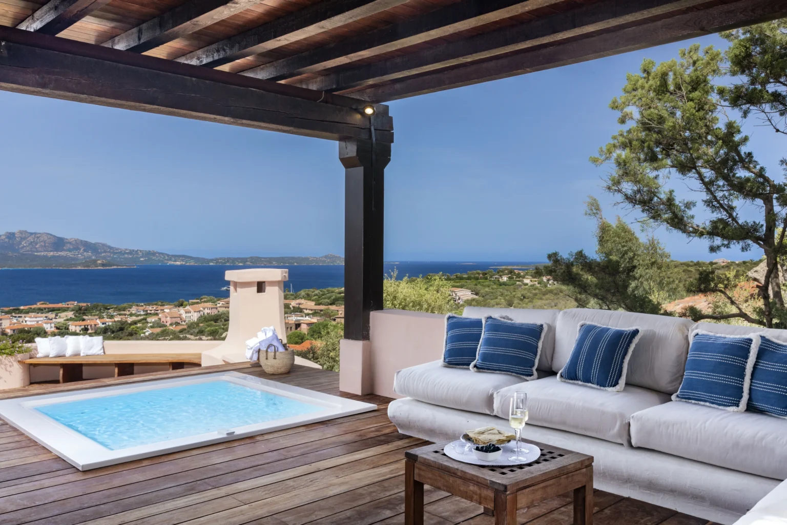 Modern outdoor living space featuring a cozy sofa with blue accent pillows, a small hot tub, and a stunning view of the ocean and landscape under a wooden pergola. Ideal for relaxation and leisure. | Only on SopranoVillas