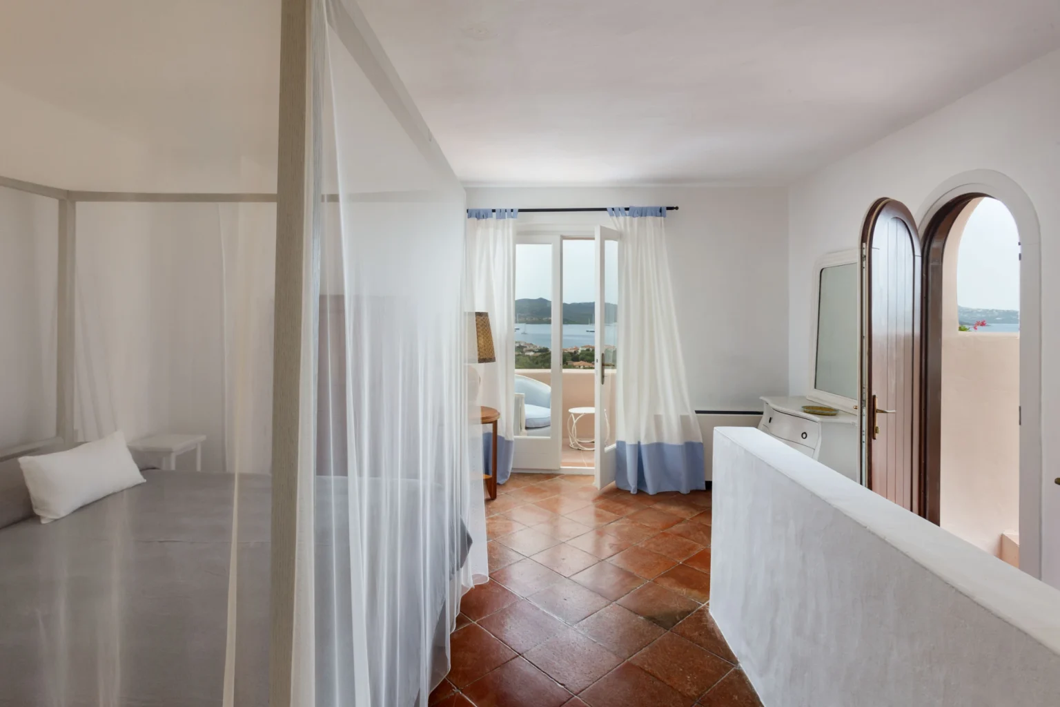 Bright and airy hotel room featuring a queen-sized bed with sheer curtains, a balcony offering scenic views, and a cozy seating area with natural light streaming in through large windows. | Only on SopranoVillas
