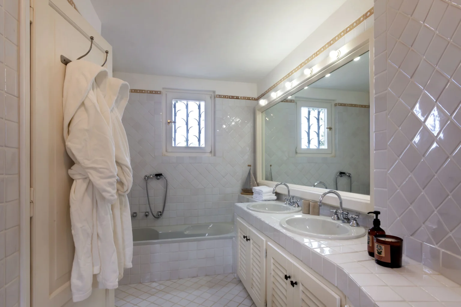 Modern bathroom featuring a white robe hanging on a hook, a large mirror with bright lighting, a bathtub, and elegant tile work. The space is well-lit, showcasing a clean and inviting atmosphere. | Only on SopranoVillas
