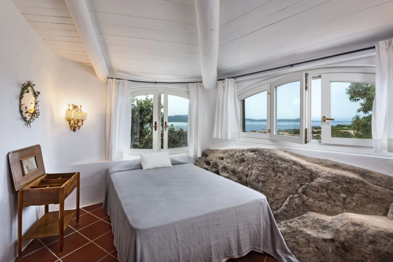 Cozy bedroom with a large bed, natural stone feature, and panoramic windows offering a view of the sea, complemented by white walls and wooden beams. | Only on SopranoVillas
