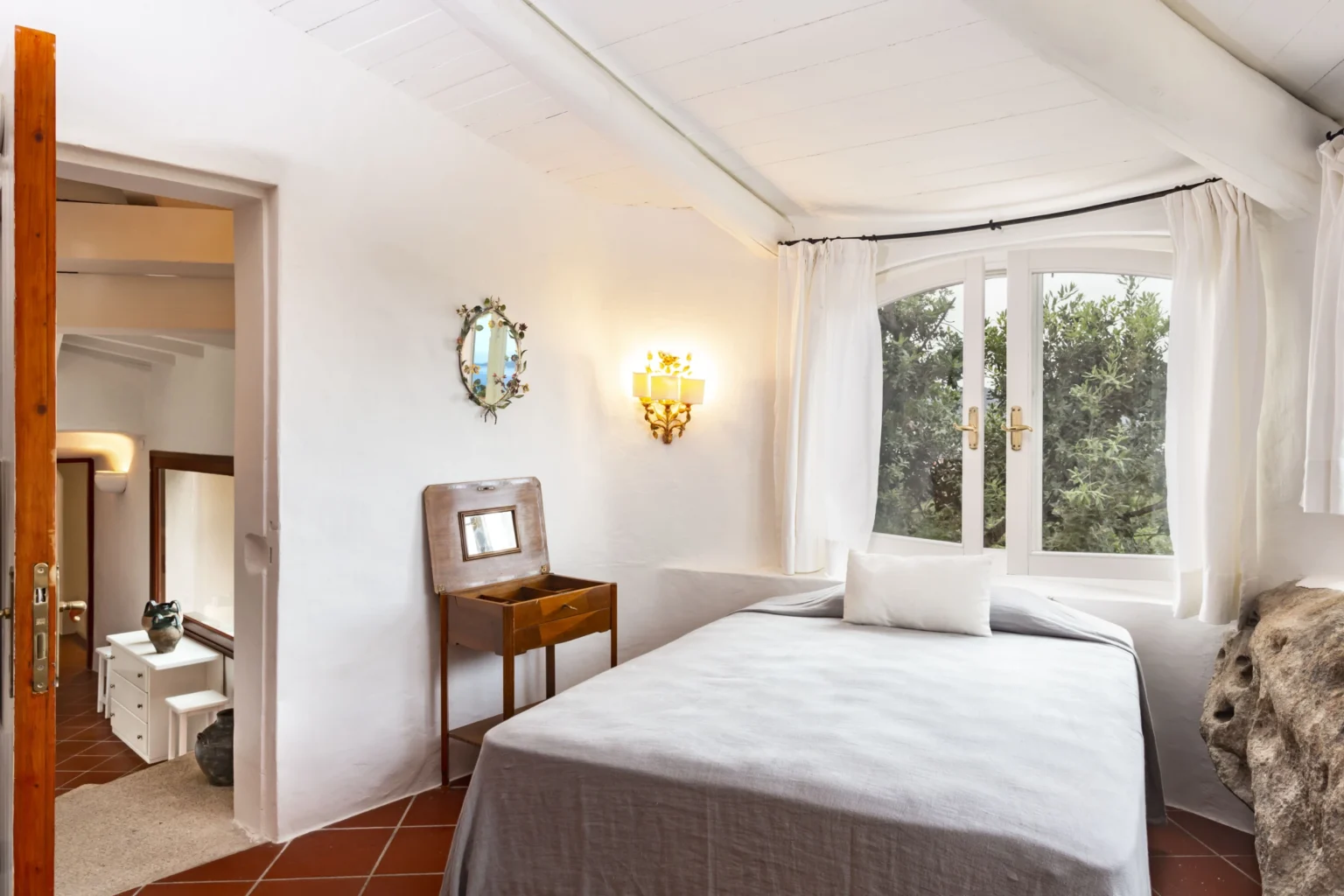 Cozy bedroom with a white bed, natural light from large windows, and decorative wall fixtures, featuring a wooden nightstand and an adjoining bathroom. | Only on SopranoVillas