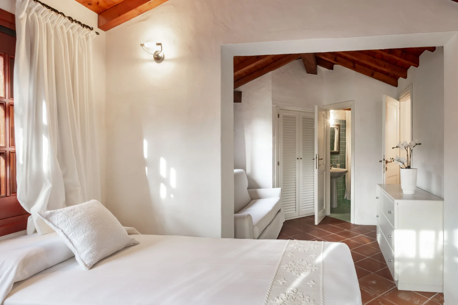 Bright and airy bedroom featuring a white bed with decorative pillows, wooden ceiling beams, and large windows draped with sheer curtains, leading to a modern bathroom and dressing area. | Only on SopranoVillas