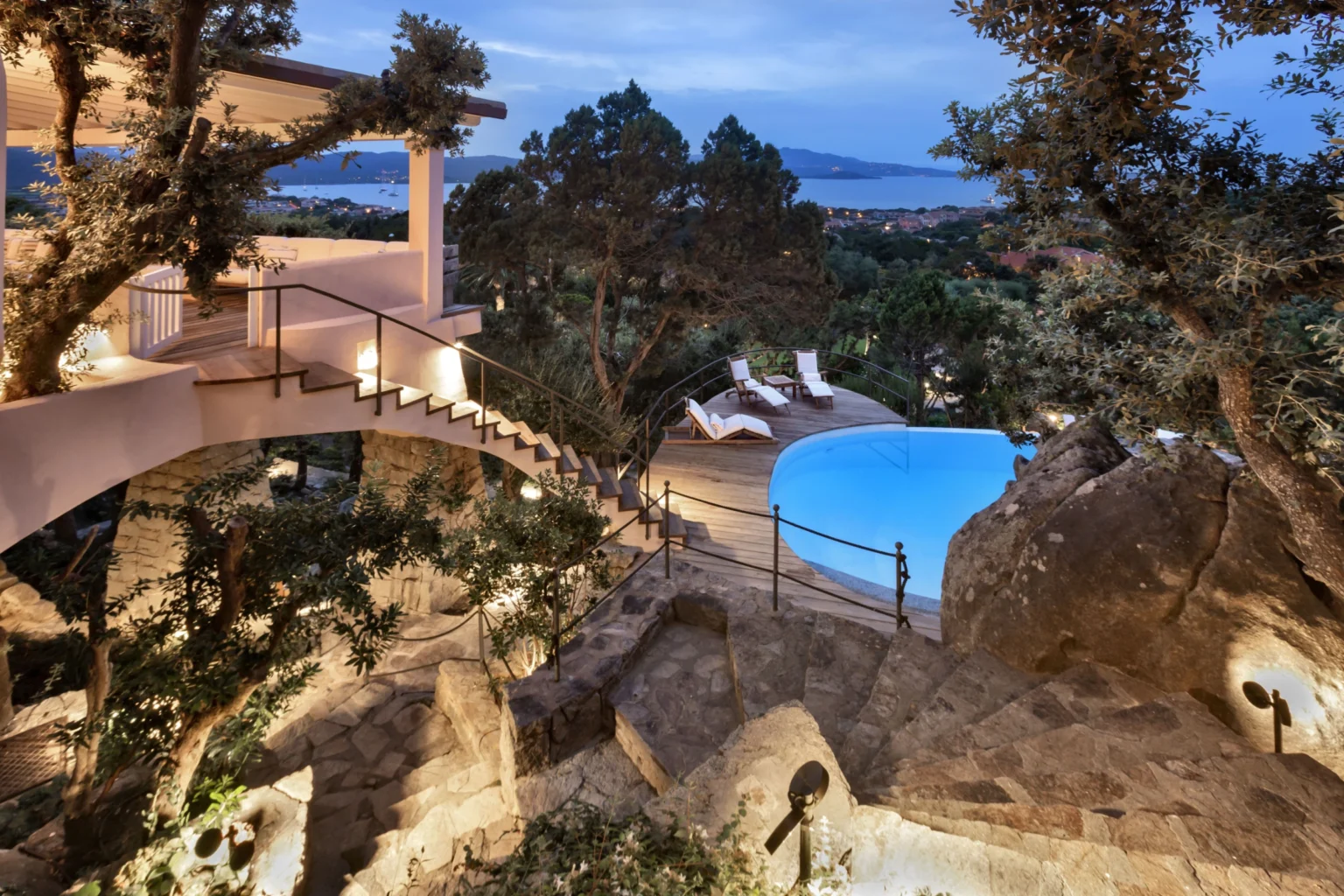 Luxurious villa with a scenic outdoor pool surrounded by lush greenery and rocky terrain, featuring elegant lounge chairs and a stunning view of the ocean at dusk. | Only on SopranoVillas