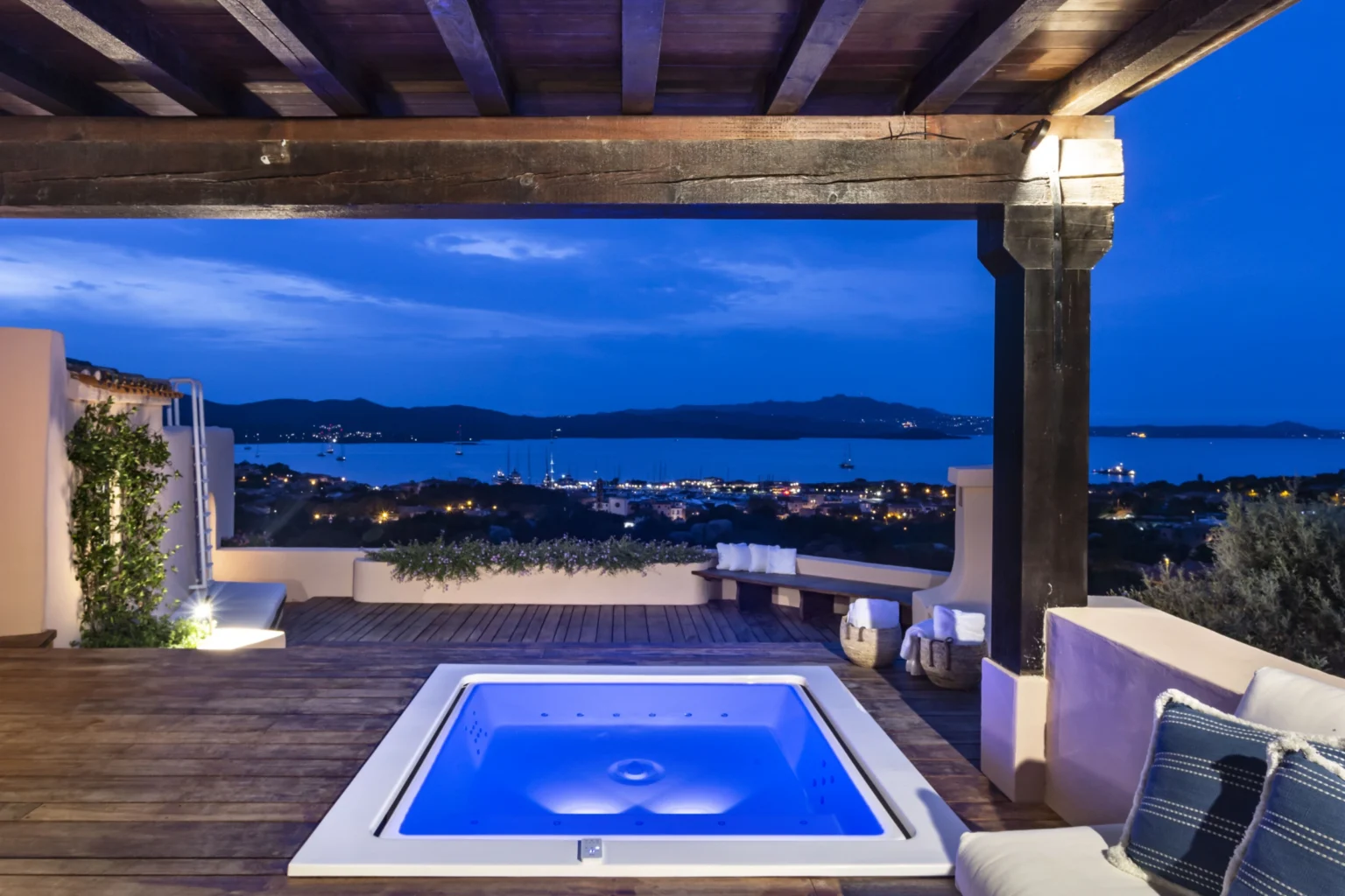Modern outdoor terrace with a hot tub overlooking a tranquil bay at night, featuring wooden beams and ambient lighting. | Only on SopranoVillas