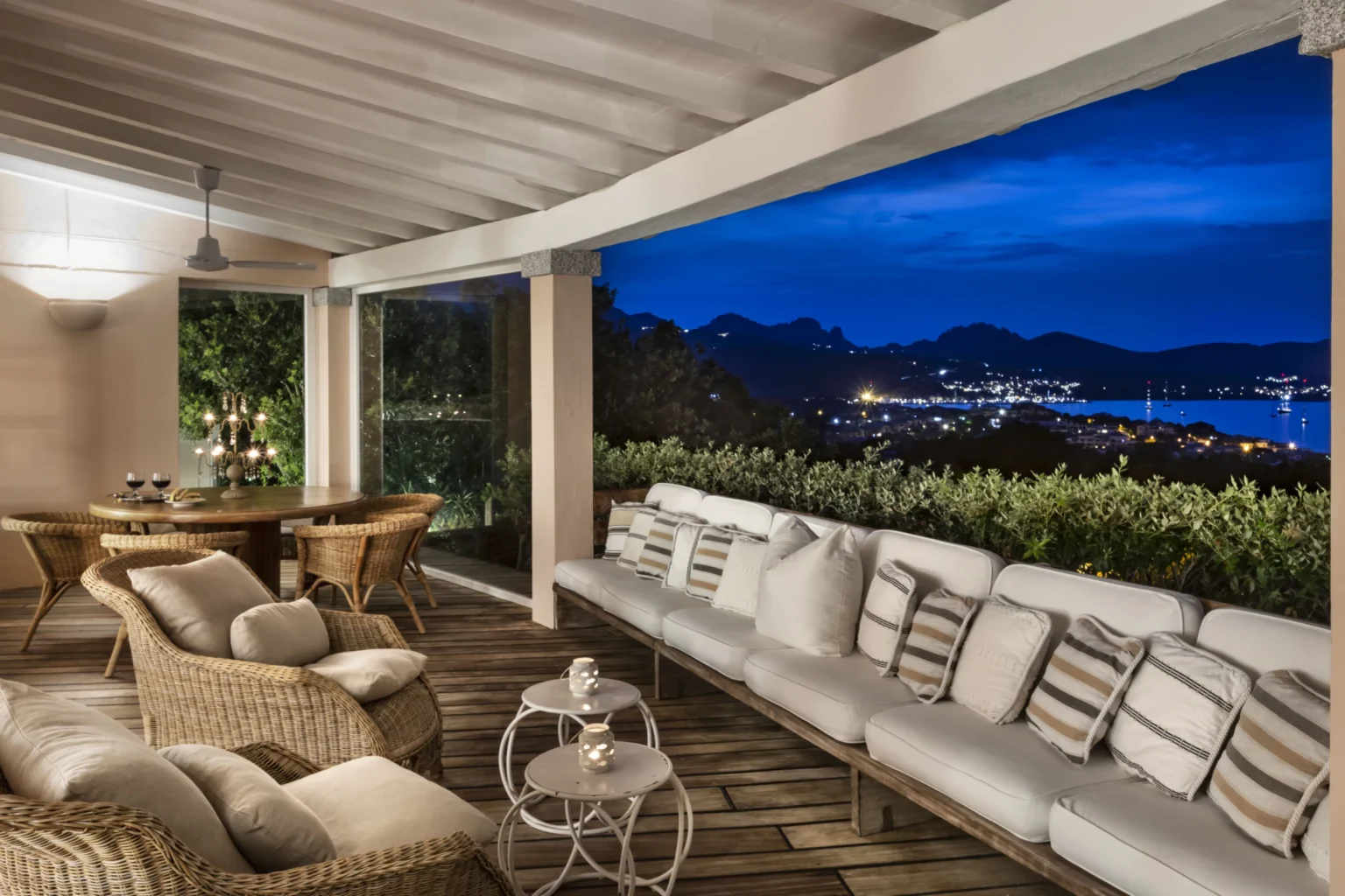Luxurious outdoor living space featuring comfortable wicker furniture, a wooden dining table, and a stunning view of a night landscape with mountains and city lights. | Only on SopranoVillas