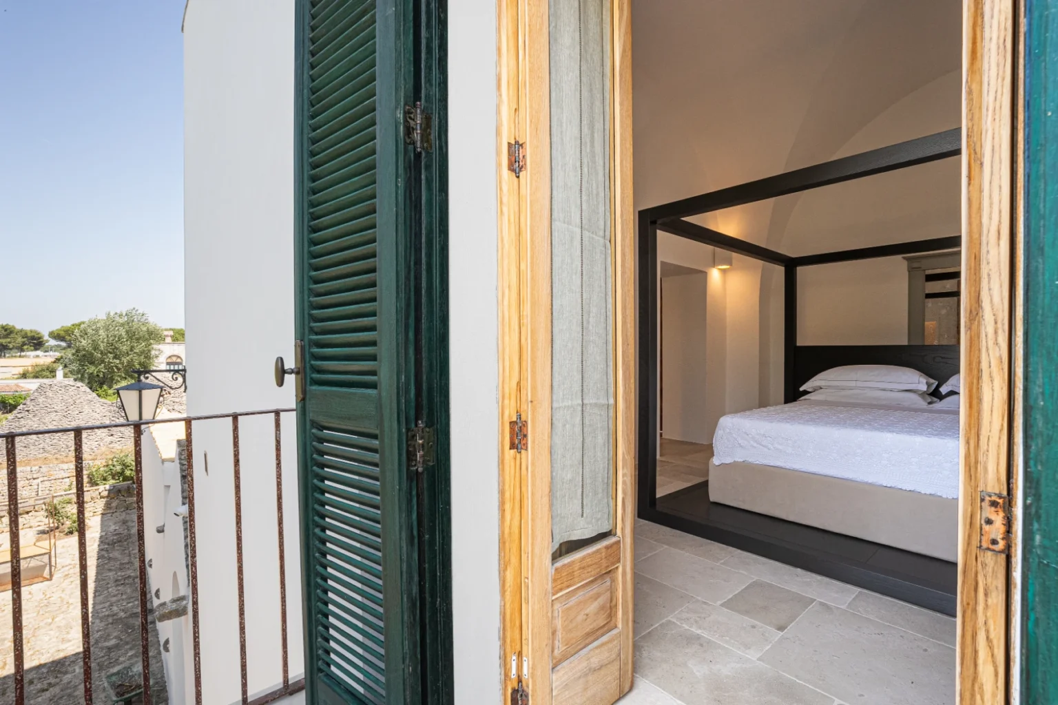 A spacious bedroom with a four-poster bed visible through an open balcony door, featuring wooden shutters and a serene outdoor view. | Only on SopranoVillas