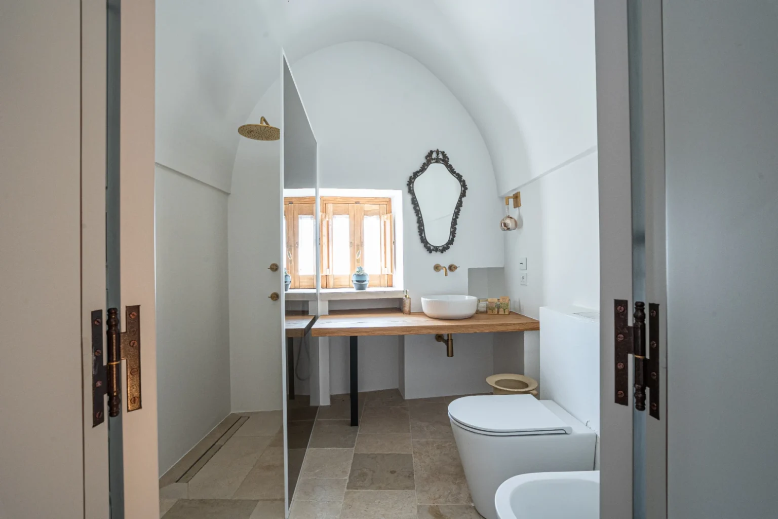 Modern bathroom interior featuring an arched ceiling, minimalist design, wooden countertop with a sink, and elegant mirror. The space includes a toilet and is illuminated by natural light from a window, creating a bright and airy atmosphere. | Only on SopranoVillas