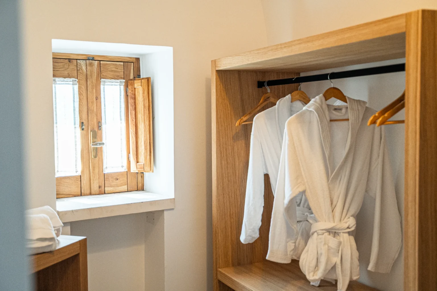 Cozy hotel room featuring a wooden wardrobe with white bathrobes hanging, complemented by a small window with wooden shutters, creating a serene and inviting atmosphere. | Only on SopranoVillas