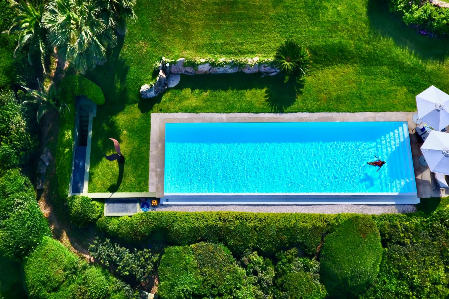 Aerial view of a luxurious swimming pool surrounded by lush greenery and palm trees, featuring lounge chairs and a serene landscape. | Only on SopranoVillas