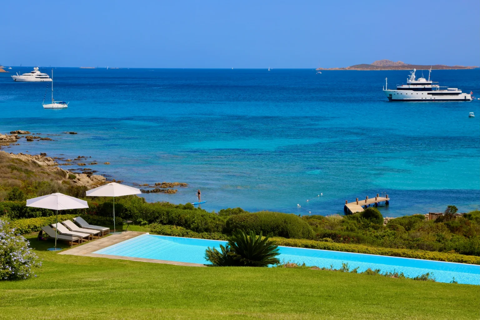 Scenic view of a luxury coastal villa with a swimming pool, lush green lawn, and yachts anchored in the turquoise sea under a clear blue sky. | Only on SopranoVillas