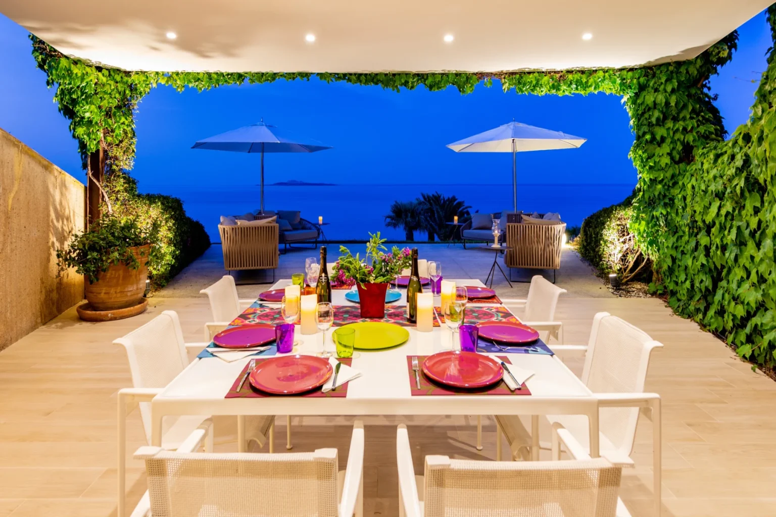 Outdoor dining area with a beautifully set table overlooking the ocean at twilight, featuring colorful plates, vibrant decor, and lush greenery. | Only on SopranoVillas