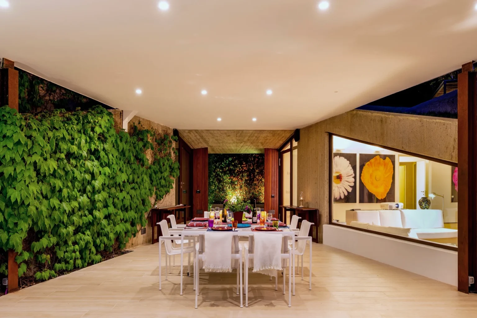 Modern outdoor dining area with a large table set for a meal, surrounded by lush greenery and contemporary architecture, featuring large windows that open to a stylish living space. | Only on SopranoVillas