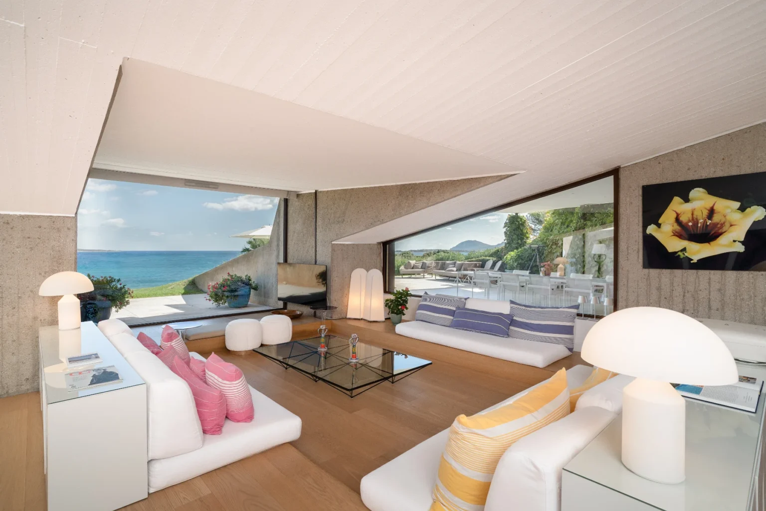 Modern coastal living room with large windows showcasing a scenic ocean view, featuring minimalist white furniture, colorful cushions, and stylish decor. | Only on SopranoVillas