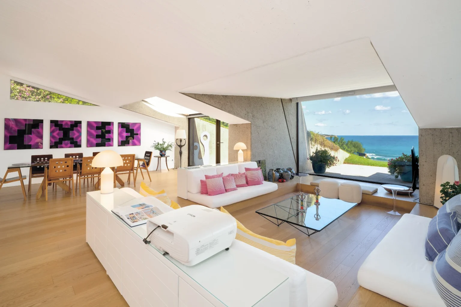 Modern living room with white furniture, large windows showcasing a coastal view, and contemporary art on the walls. The space features wooden flooring and a bright, airy atmosphere, ideal for relaxation and entertaining. | Only on SopranoVillas