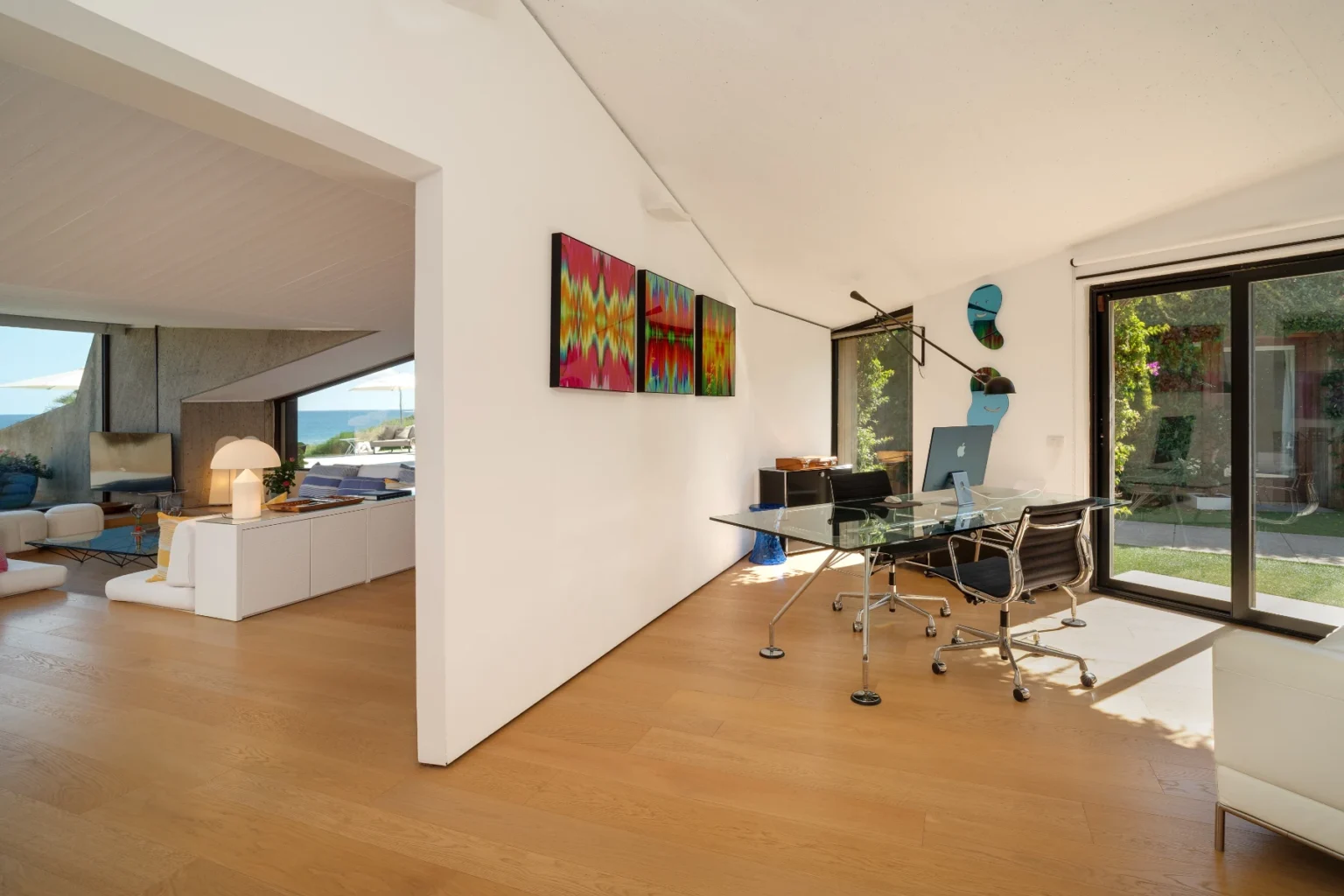 Modern home office with glass desk and ergonomic chair, featuring colorful artwork on the walls and large windows that provide natural light and a view of the outdoor space. | Only on SopranoVillas