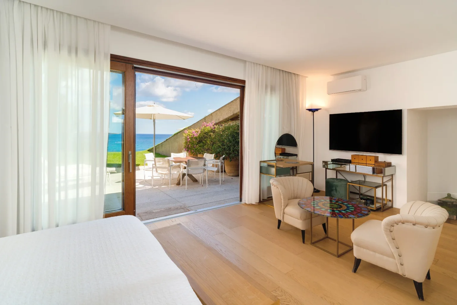 Cozy hotel room with modern decor featuring a large window that opens to a patio with ocean views, showcasing comfortable seating and a stylish coffee table. | Only on SopranoVillas
