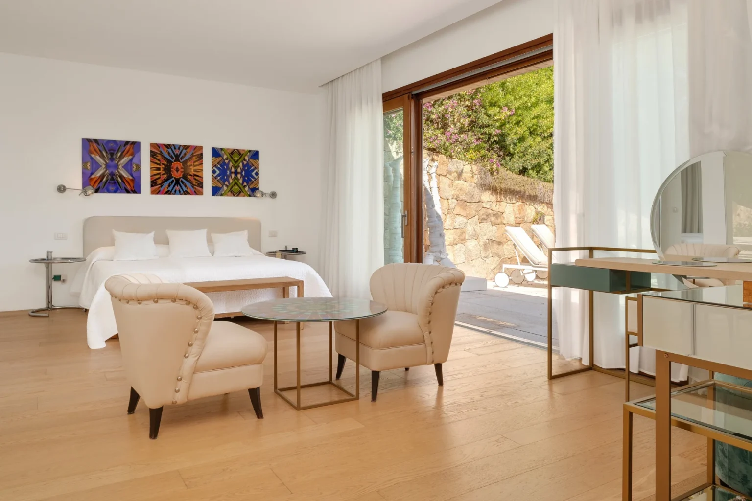 Modern hotel room featuring a king-sized bed, stylish beige armchairs, a glass table, and large windows that open to a scenic outdoor view, decorated with colorful artwork on the walls. | Only on SopranoVillas