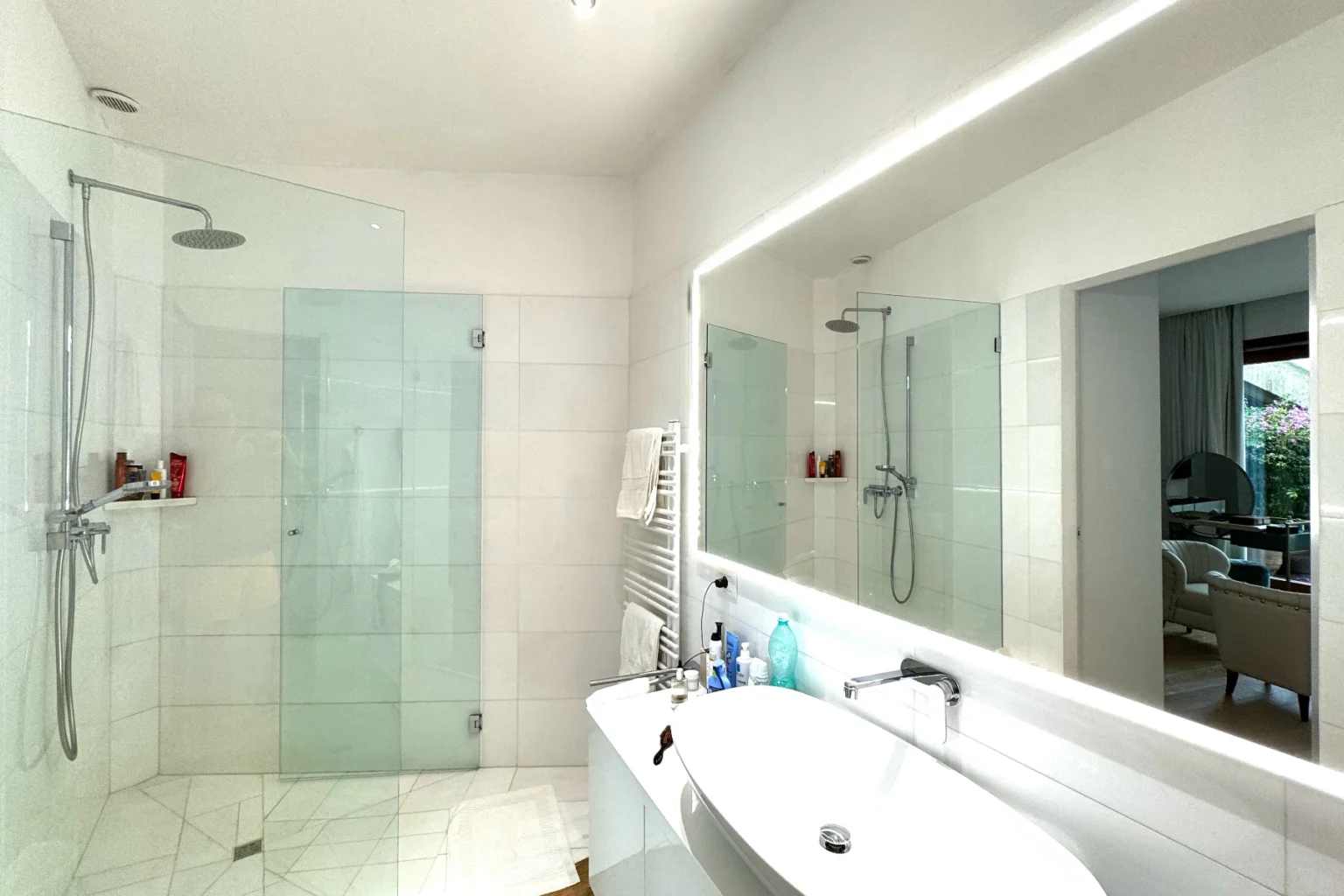 Modern bathroom featuring a spacious walk-in shower with glass doors, a sleek white vanity, and a large mirror reflecting a bright and airy design. The room is illuminated by recessed lighting, enhancing the contemporary aesthetic. | Only on SopranoVillas