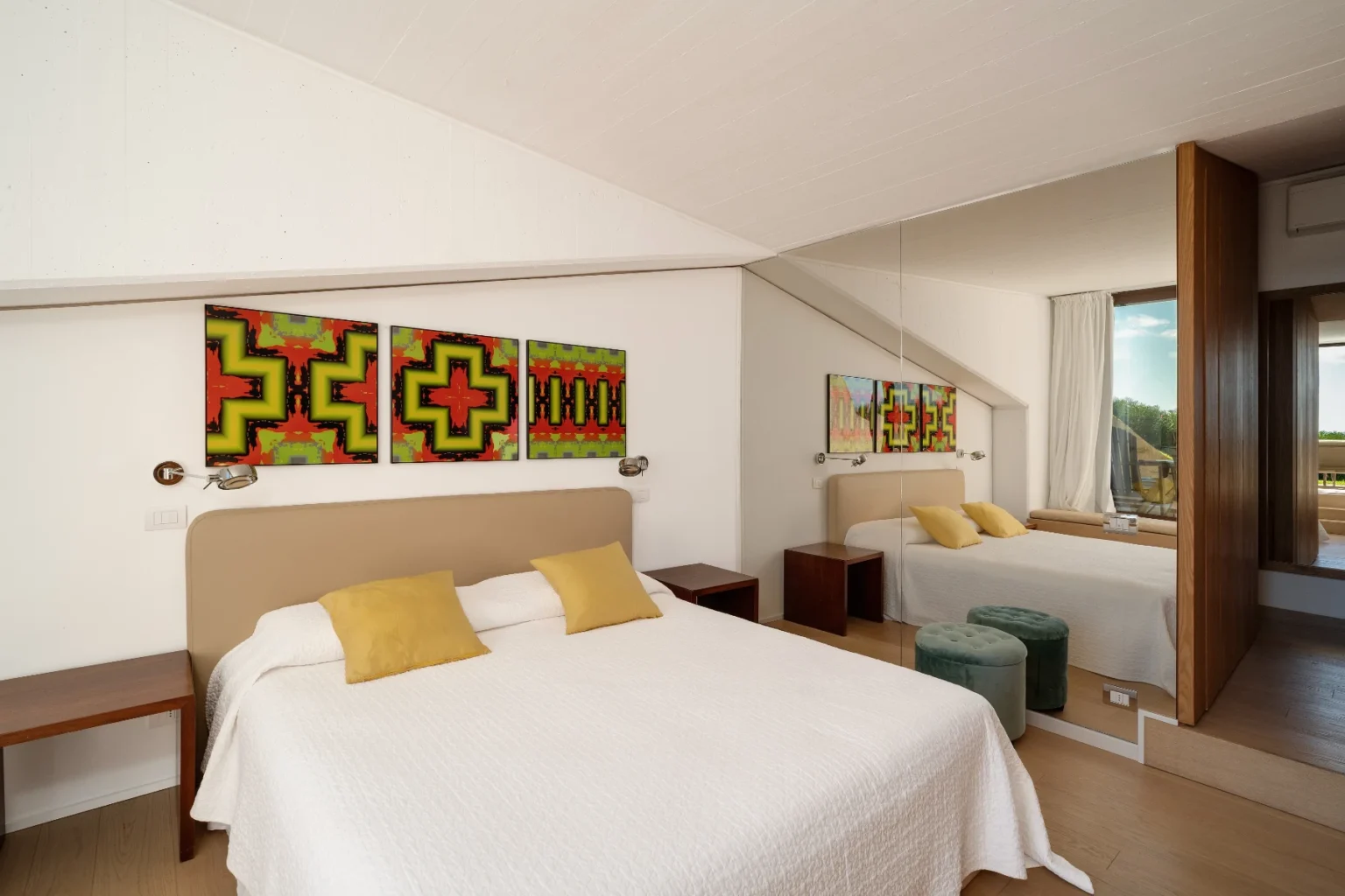 Modern hotel room featuring a double bed with white linens and yellow accent pillows, complemented by colorful artwork on the walls. The room includes a wooden nightstand and green ottomans, with large windows allowing natural light to brighten the space. | Only on SopranoVillas