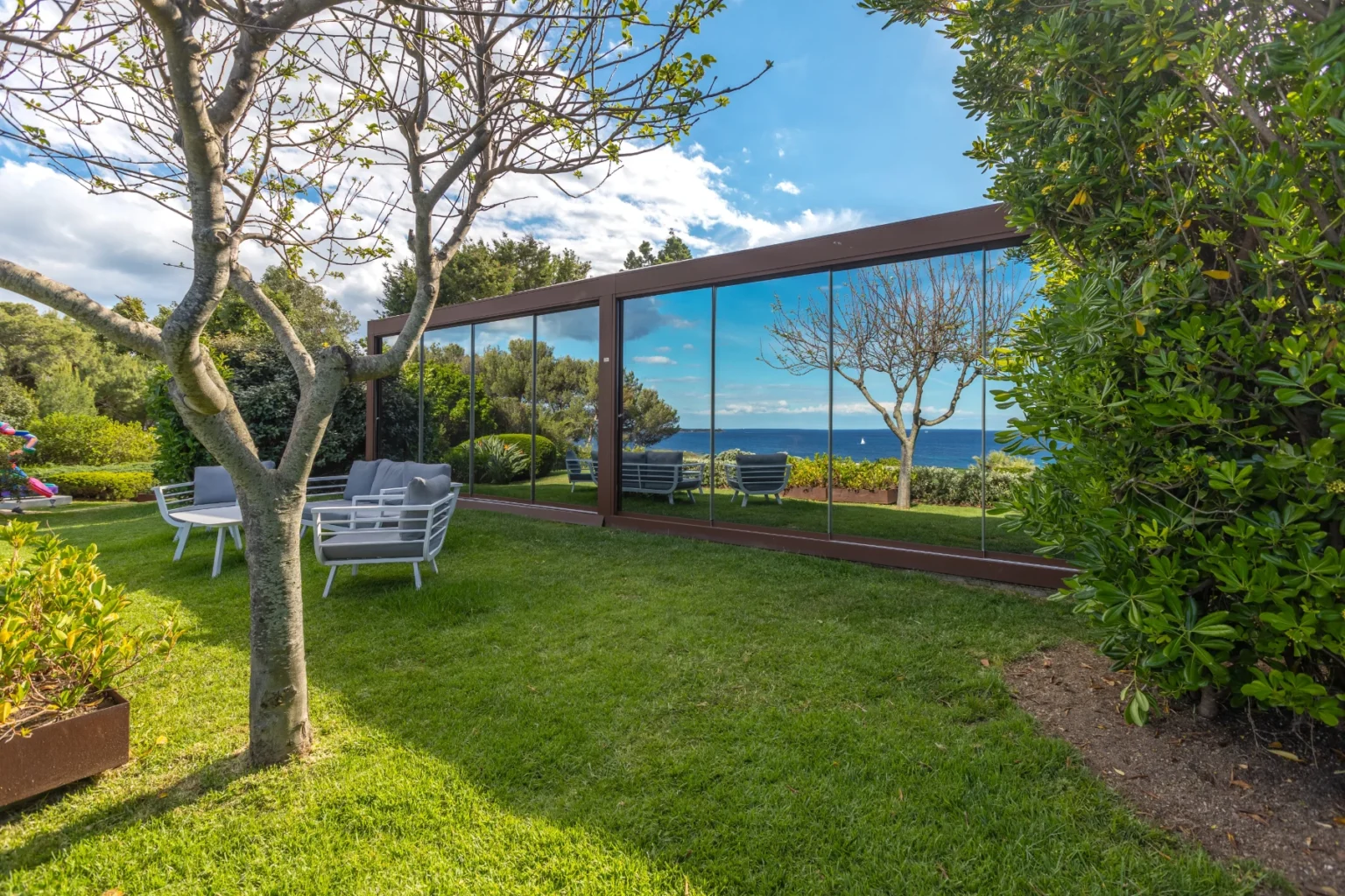 Modern garden with lush greenery, a glass wall reflecting the ocean view, and outdoor seating in a serene landscape. | Only on SopranoVillas