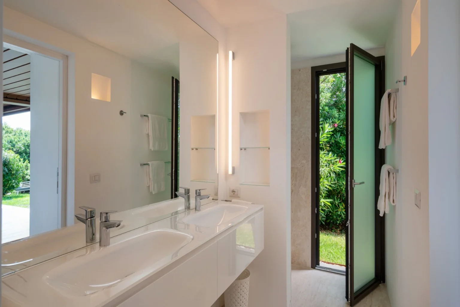 Modern bathroom interior featuring a double sink vanity, sleek fixtures, and a large window with a view of greenery. The design emphasizes natural light and minimalist aesthetics, creating a serene and inviting atmosphere. | Only on SopranoVillas