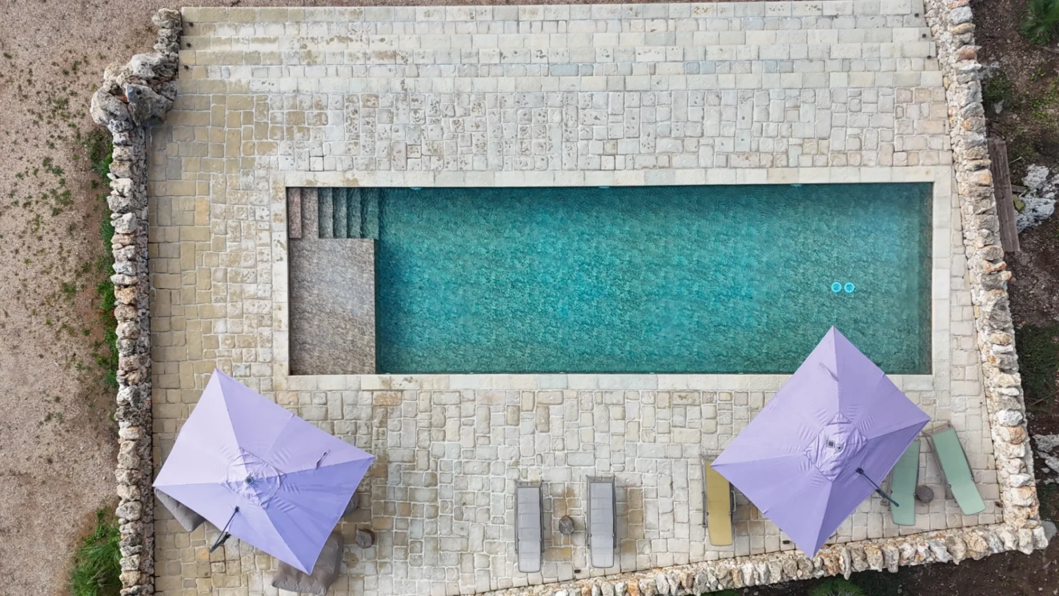Aerial view of a serene swimming pool surrounded by stone flooring, featuring two purple umbrellas and lounge chairs, ideal for relaxation in a luxury outdoor setting. | Only on SopranoVillas