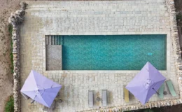 Aerial view of a serene swimming pool surrounded by stone flooring, featuring two purple umbrellas and lounge chairs, ideal for relaxation in a luxury outdoor setting.