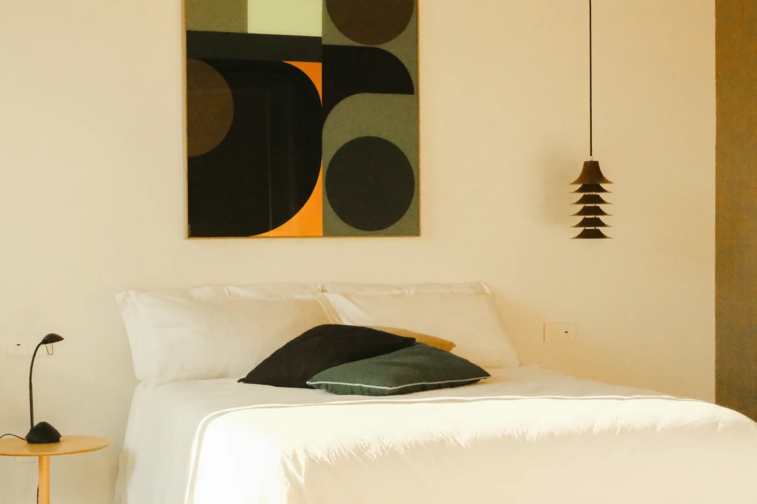 Modern bedroom featuring a minimalist design with a white bed, decorative pillows, a stylish pendant light, and an abstract wall art piece with geometric shapes in muted colors. | Only on SopranoVillas