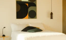 Modern bedroom featuring a minimalist design with a white bed, decorative pillows, a stylish pendant light, and an abstract wall art piece with geometric shapes in muted colors.