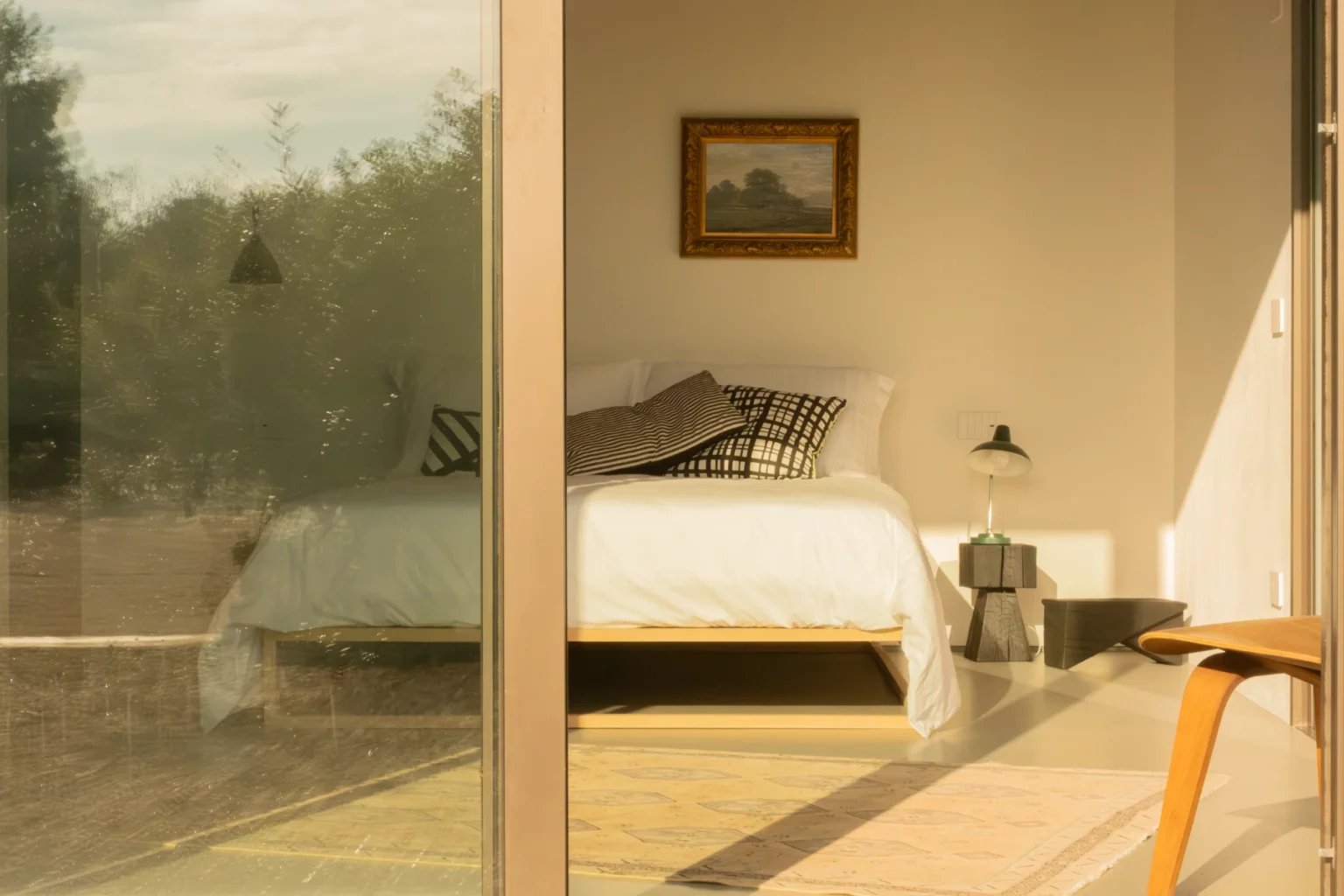 Cozy bedroom with a large window showcasing a serene view, featuring a bed with white linens, decorative pillows, a bedside lamp, and a small table. The warm lighting highlights the minimalist decor and a framed painting on the wall. | Only on SopranoVillas