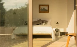 Cozy bedroom with a large window showcasing a serene view, featuring a bed with white linens, decorative pillows, a bedside lamp, and a small table. The warm lighting highlights the minimalist decor and a framed painting on the wall.