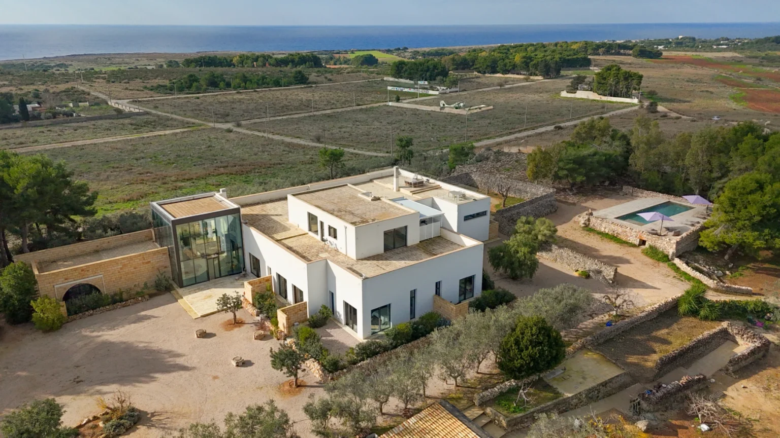 Aerial view of a modern white villa surrounded by lush greenery and open fields, with a swimming pool and ocean in the background. The property features landscaped gardens and a contemporary design, ideal for luxury living. | Only on SopranoVillas
