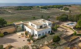 Aerial view of a modern white villa surrounded by lush greenery and open fields, with a swimming pool and ocean in the background. The property features landscaped gardens and a contemporary design, ideal for luxury living.
