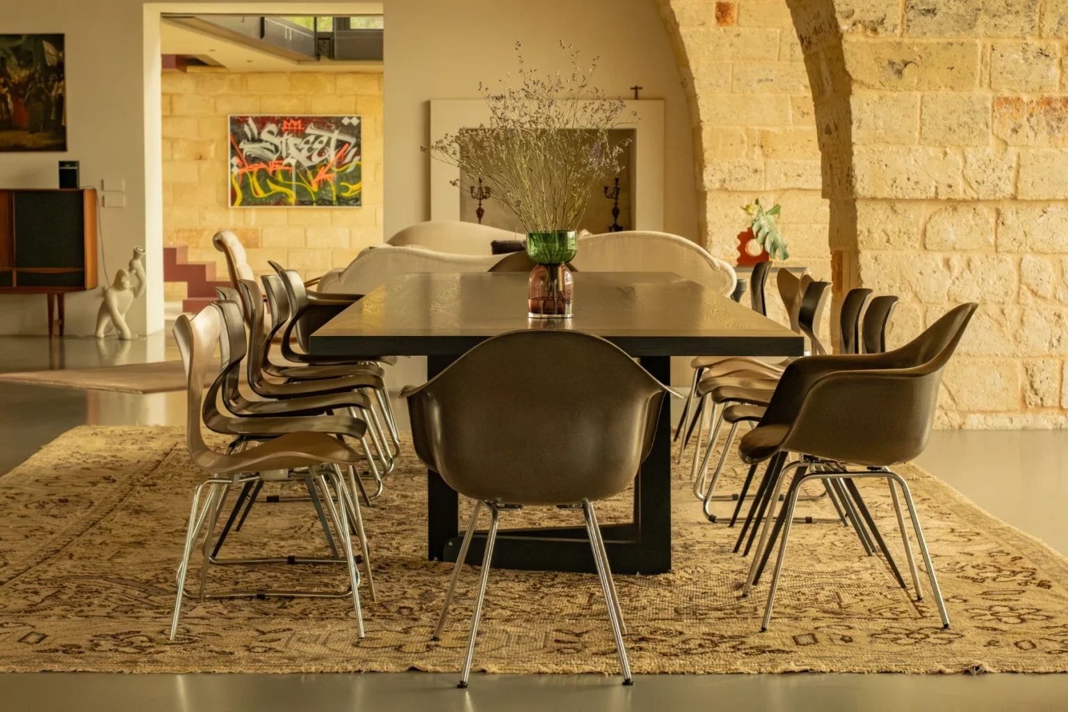Modern dining room featuring a large black table surrounded by stylish brown chairs, set on a decorative rug. The space is illuminated with natural light and decorated with flowers and artwork, creating an inviting atmosphere for gatherings. | Only on SopranoVillas