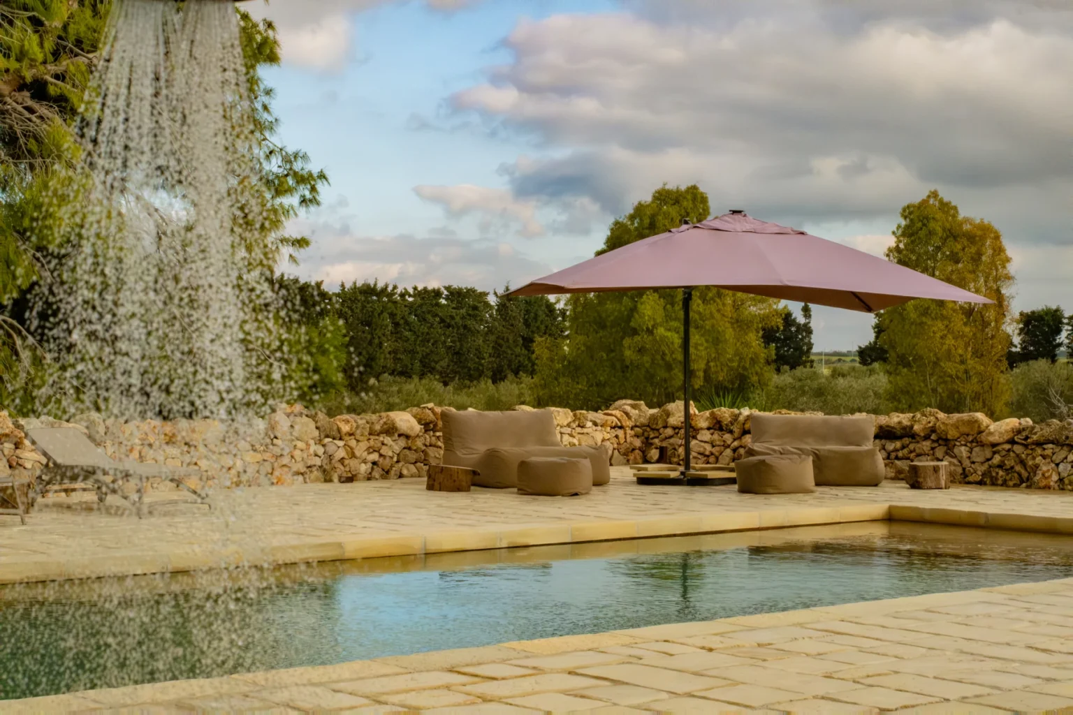 Serene outdoor pool area featuring a gentle waterfall, comfortable seating, and a pink umbrella, surrounded by lush greenery and a stone wall. Ideal for relaxation and leisure. | Only on SopranoVillas