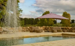 Serene outdoor pool area featuring a gentle waterfall, comfortable seating, and a pink umbrella, surrounded by lush greenery and a stone wall. Ideal for relaxation and leisure.