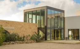 Modern architectural design featuring a glass and stone house with a spacious outdoor area, surrounded by greenery under a cloudy sky.