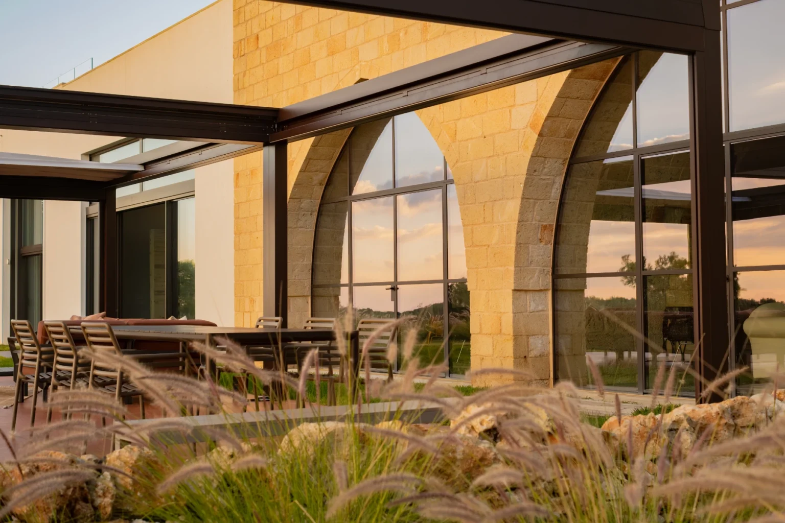 Modern outdoor seating area with a stone and glass structure, featuring stylish furniture and lush landscaping, perfect for dining or relaxation. | Only on SopranoVillas