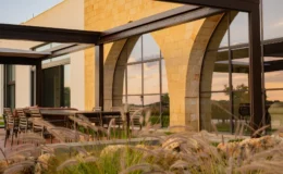 Modern outdoor seating area with a stone and glass structure, featuring stylish furniture and lush landscaping, perfect for dining or relaxation.