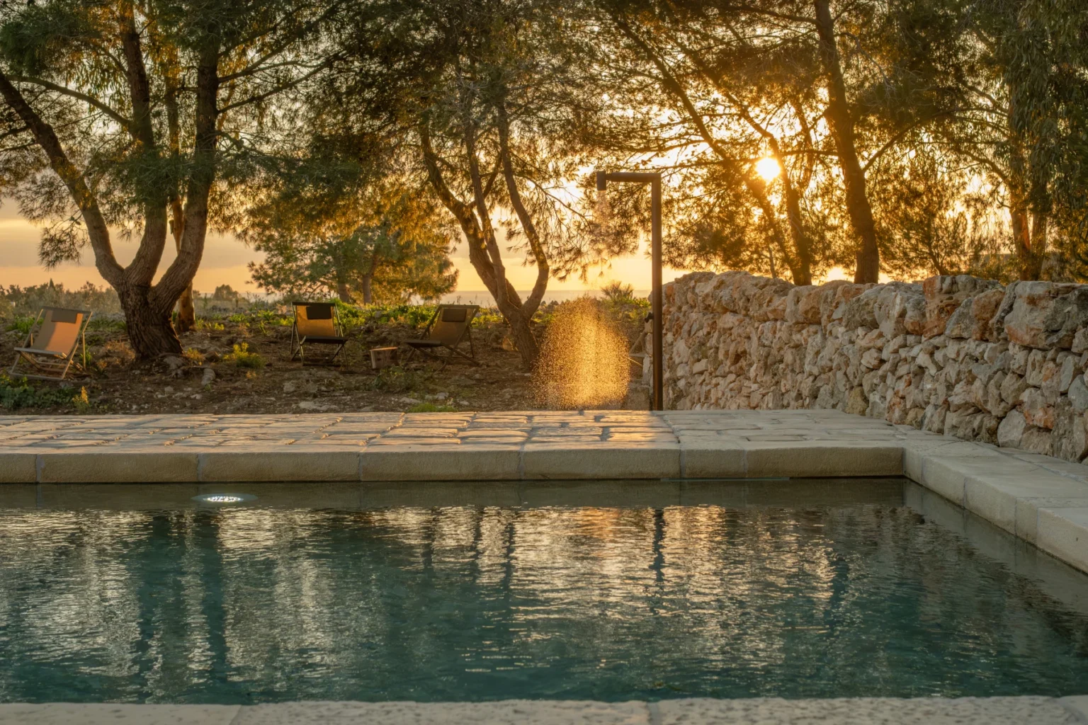 Serene outdoor swimming pool surrounded by lush trees at sunset, with warm golden light reflecting on the water's surface and a stone wall in the background. | Only on SopranoVillas