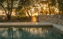 Serene outdoor swimming pool surrounded by lush trees at sunset, with warm golden light reflecting on the water's surface and a stone wall in the background.
