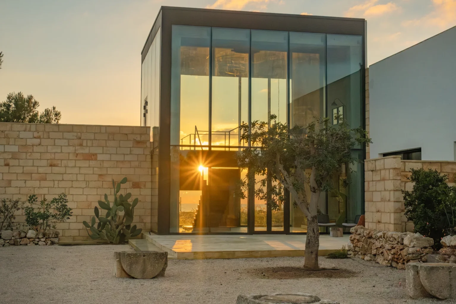 Modern glass house at sunset surrounded by natural landscaping, featuring a stone wall and sculptures in the foreground, emphasizing contemporary architecture and serene outdoor living. | Only on SopranoVillas