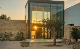 Modern glass house at sunset surrounded by natural landscaping, featuring a stone wall and sculptures in the foreground, emphasizing contemporary architecture and serene outdoor living.